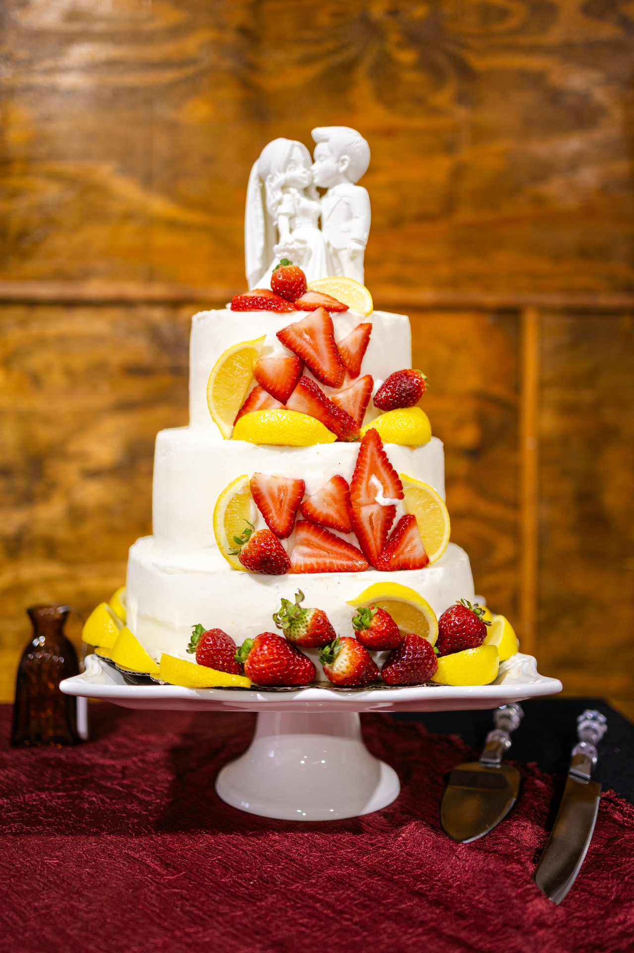 Three-tier white wedding cake topped with a porcelain bride and groom, decorated with fresh strawberries and lemon slices